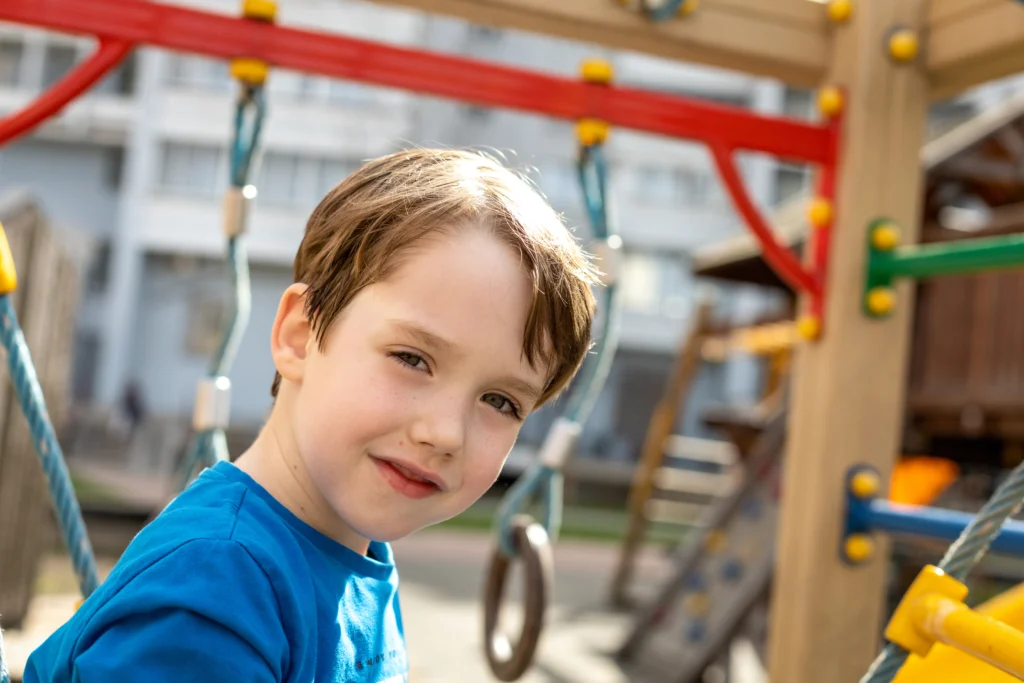 The image shows a child on a playground. The child's face is blurred out, and the child is wearing a blue shirt. The playground equipment includes ropes and wooden structures, with some colorful elements such as red and yellow bars. There are buildings in the background.